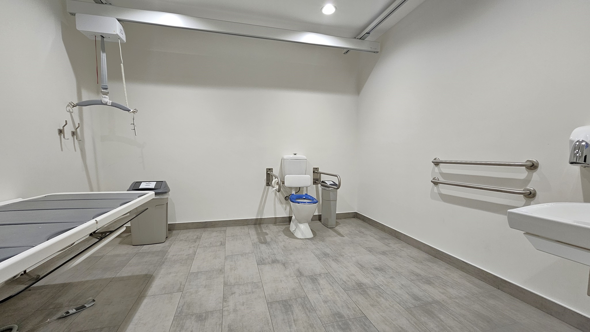 On the left, there is a grey changing table attached to the wall, with a bin underneath it. In the middle, there is a white toilet with a blue seat cover, a small bin next to it, and a metal grab bar on the wall. On the right, there is a white sink with a soap dispenser above it and two metal grab bars on the wall. The floor is covered with large grey tiles, and the ceiling is white with recessed lighting and exposed metal beams.