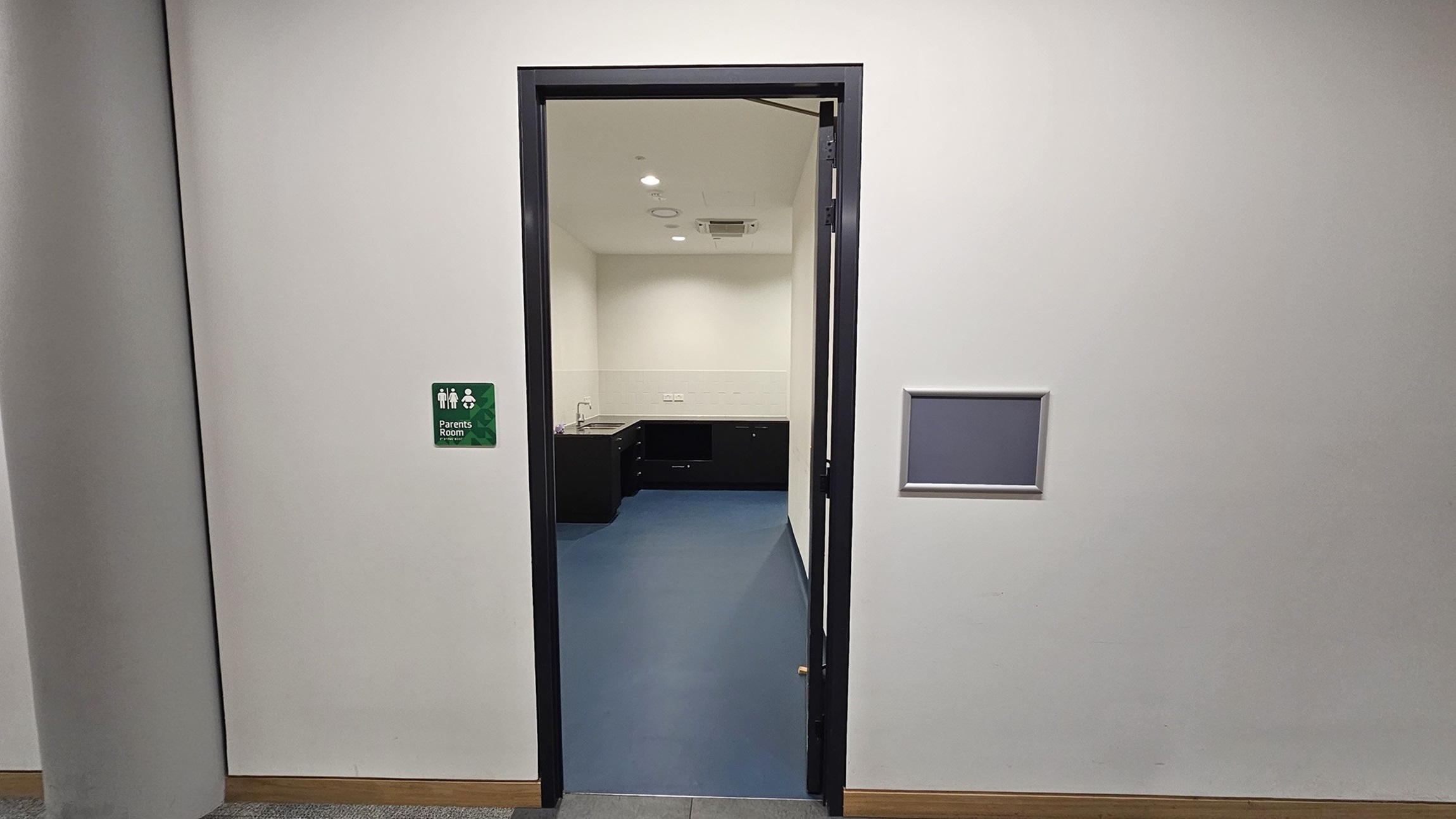 On the left, there is a cylindrical column and a green sign indicating "Parents Room" with a white icon. In the middle, there is an open doorway with a black frame leading to a room with a blue floor, white walls, and black cabinets. On the right, there is a rectangular recessed panel in the wall. The floor outside the room is covered with grey tiles and a wooden baseboard. The ceiling is white with recessed lighting.