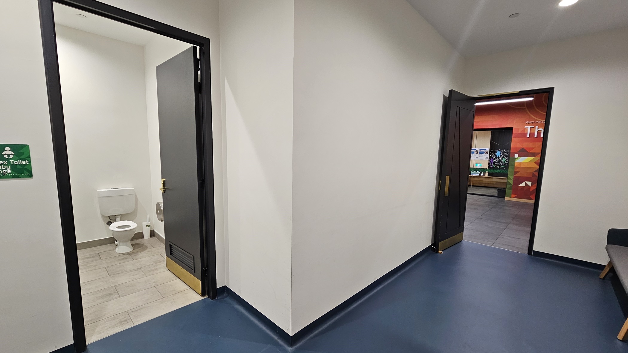 On the left, there is an open door with a black frame leading to a toilet with a white toilet bowl and grey floor tiles. In the middle, there is a white wall with a corner. On the right, there is another open door with a black frame leading to a room with colourful wall art and a display board. The floor in the foreground is covered with blue linoleum, and the ceiling is white with recessed lighting.