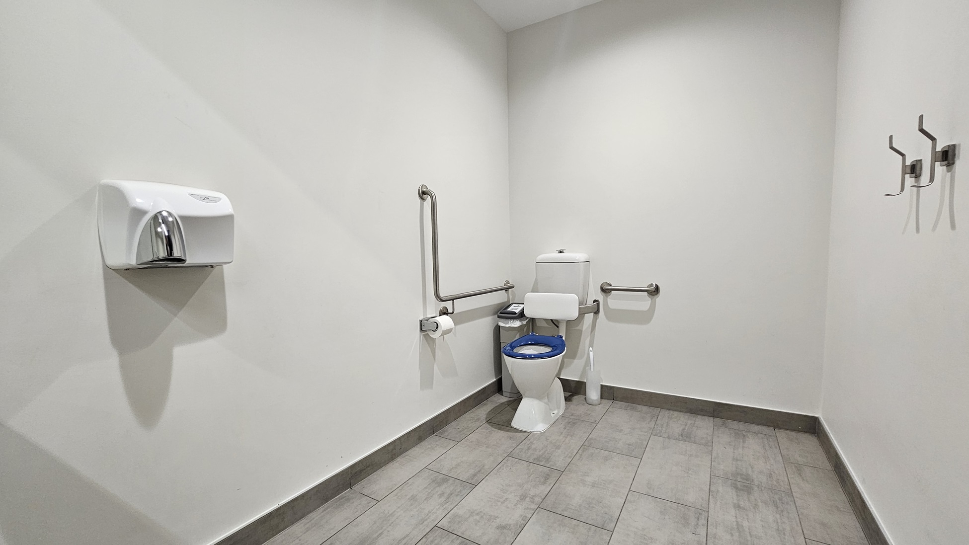 On the left, there is a white wall-mounted paper towel dispenser. In the middle, there is a white toilet with a blue seat, flanked by metal grab bars and a toilet paper holder. On the right, there are three metal coat hooks attached to the wall. The floor is covered with large grey tiles, and the ceiling is white with a slight slope.