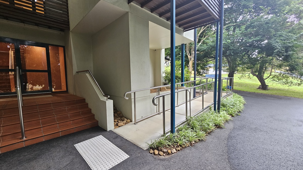 On the left, there is a set of brown tiled steps leading to a glass door. A metal handrail runs alongside the steps. In the centre, a ramp with metal railings provides an accessible path, bordered by small plants and rocks. On the right, a paved path curves away, with trees and greenery in the background. The ceiling above the ramp is supported by blue metal beams. On the left, there is a set of brown tiled steps leading to a glass door. A metal handrail runs alongside the steps. In the centre, a ramp with metal railings provides an accessible path, bordered by small plants and rocks. On the right, a paved path curves away, with trees and greenery in the background. The ceiling above the ramp is supported by blue metal beams.