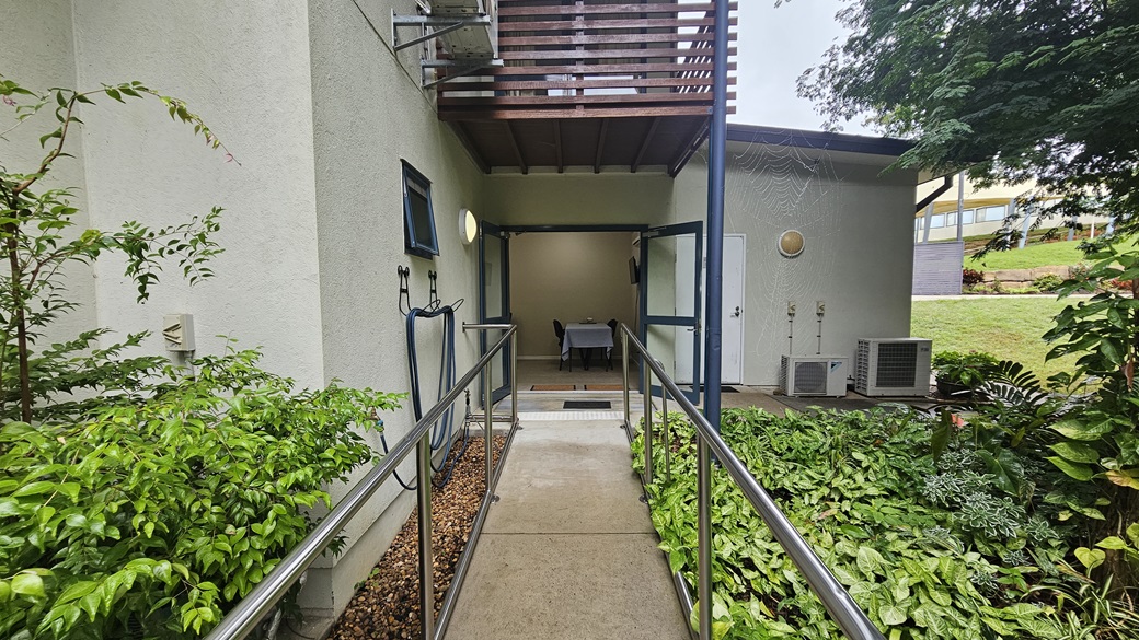 A narrow concrete path with metal railings leads to a building entrance. On the left, there are lush green plants and a small window on the building wall. On the right, more greenery is visible, along with air conditioning units and a door. Above the entrance, there is a wooden balcony. The background shows a grassy area with trees and a glimpse of another building. A narrow concrete path with metal railings leads to a building entrance. On the left, there are lush green plants and a small window on the building wall. On the right, more greenery is visible, along with air conditioning units and a door. Above the entrance, there is a wooden balcony. The background shows a grassy area with trees and a glimpse of another building.