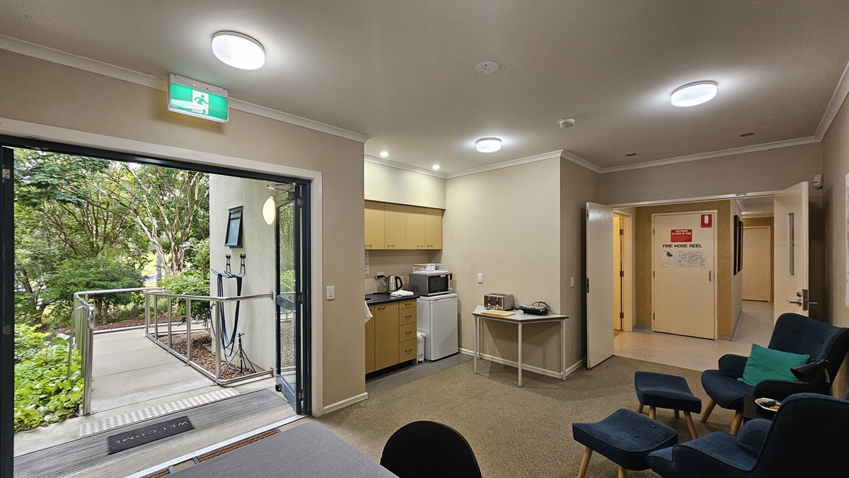 A room with beige walls and a carpeted floor. On the left, a glass door opens to a garden with trees and a ramp. In the middle, a kitchenette with wooden cabinets, a microwave, kettle, and mini-fridge. On the right, a hallway with open doors and a red emergency sign. Several dark blue chairs with teal cushions are arranged in the foreground. The ceiling has recessed lighting and a glowing exit sign above the door. A room with beige walls and a carpeted floor. On the left, a glass door opens to a garden with trees and a ramp. In the middle, a kitchenette with wooden cabinets, a microwave, kettle, and mini-fridge. On the right, a hallway with open doors and a red emergency sign. Several dark blue chairs with teal cushions are arranged in the foreground. The ceiling has recessed lighting and a glowing exit sign above the door.