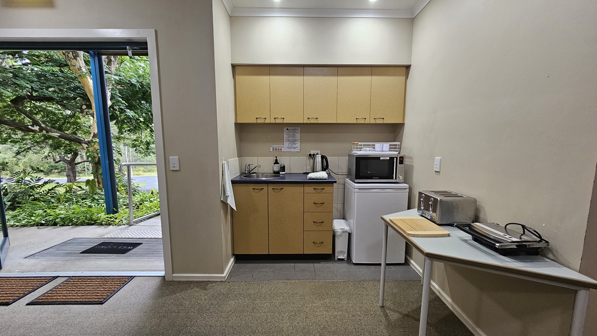 A small kitchen area with light wood cabinets and a black countertop. On the counter are a kettle and a microwave. Below is a mini fridge. To the right, a table holds a toaster and a sandwich press. The floor is carpeted, and the ceiling has recessed lighting. On the left, an open door leads outside, revealing lush greenery and a pathway. A small kitchen area with light wood cabinets and a black countertop. On the counter are a kettle and a microwave. Below is a mini fridge. To the right, a table holds a toaster and a sandwich press. The floor is carpeted, and the ceiling has recessed lighting. On the left, an open door leads outside, revealing lush greenery and a pathway.