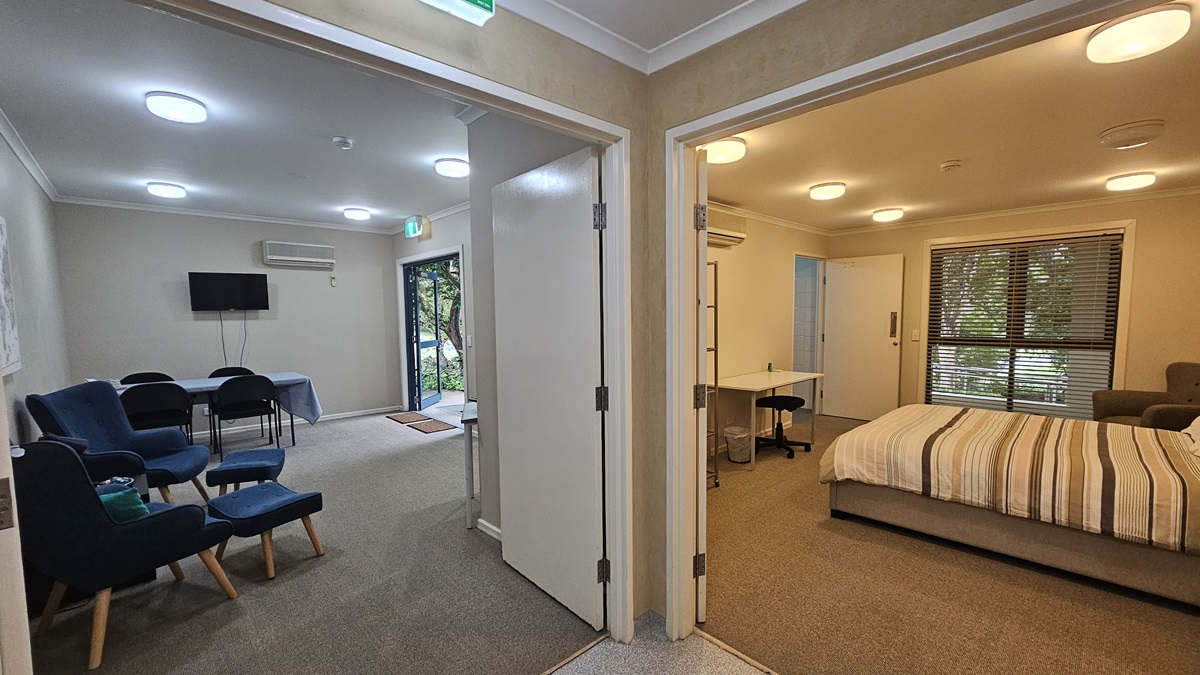 A view of two rooms from a hallway. On the left, a lounge area with blue chairs, a small table, a wall-mounted TV, and a glass door leading outside. On the right, a bedroom with a striped bed, a desk with a chair, and a window with blinds. Both rooms have beige walls and carpeted floors. Ceiling lights are visible in both spaces. A view of two rooms from a hallway. On the left, a lounge area with blue chairs, a small table, a wall-mounted TV, and a glass door leading outside. On the right, a bedroom with a striped bed, a desk with a chair, and a window with blinds. Both rooms have beige walls and carpeted floors. Ceiling lights are visible in both spaces.