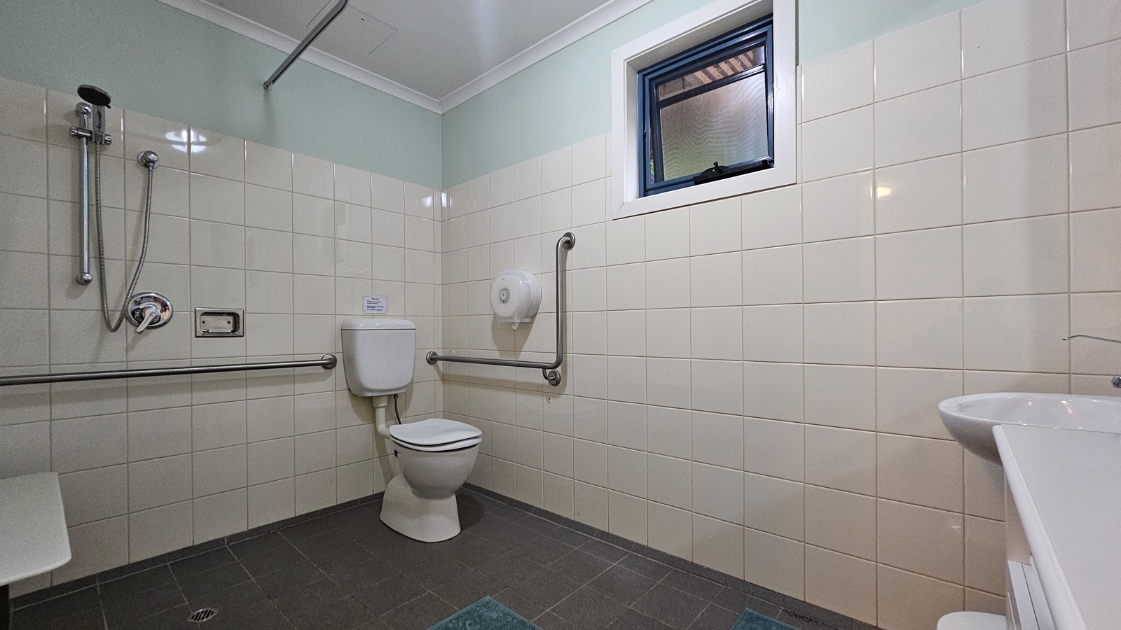 A bathroom with light-coloured tiled walls and a dark tiled floor. On the left, there is a shower with a handheld showerhead and a horizontal grab bar. In the centre, a toilet with a vertical grab bar beside it. Above the toilet, a small window with a blue frame. On the right, a white sink with a tap. The ceiling is white with a simple design. A bathroom with light-coloured tiled walls and a dark tiled floor. On the left, there is a shower with a handheld showerhead and a horizontal grab bar. In the centre, a toilet with a vertical grab bar beside it. Above the toilet, a small window with a blue frame. On the right, a white sink with a tap. The ceiling is white with a simple design.