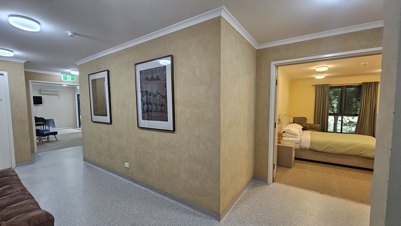 A hallway with light-coloured walls and a speckled floor. On the left wall, two framed artworks are displayed. To the right, an open doorway leads to a bedroom with a bed, bedside table, and a window with curtains. In the background, another room is visible with chairs and a table, and an exit sign above a door. The ceiling has recessed circular lights. A hallway with light-coloured walls and a speckled floor. On the left wall, two framed artworks are displayed. To the right, an open doorway leads to a bedroom with a bed, bedside table, and a window with curtains. In the background, another room is visible with chairs and a table, and an exit sign above a door. The ceiling has recessed circular lights.