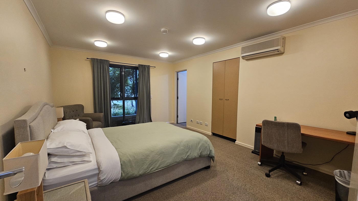 A bedroom with a bed on the left, covered with a green duvet and white pillows. Next to the bed is a grey armchair. A window with green curtains is in the background. On the right, there's a wooden desk with a grey office chair and a small bin underneath. Above the desk is an air conditioning unit. A wardrobe with two doors is next to the desk. The ceiling has several round lights, and the floor is carpeted. A bedroom with a bed on the left, covered with a green duvet and white pillows. Next to the bed is a grey armchair. A window with green curtains is in the background. On the right, there's a wooden desk with a grey office chair and a small bin underneath. Above the desk is an air conditioning unit. A wardrobe with two doors is next to the desk. The ceiling has several round lights, and the floor is carpeted.