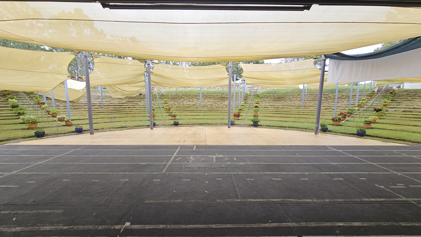 An outdoor amphitheatre with tiered grass seating in the background, arranged in a semi-circle. The foreground features a dark, paved stage area with visible lines. Overhead, beige fabric canopies are stretched across metal poles, providing shade. Potted plants with flowers are placed along the steps and near the poles, adding greenery to the scene. An outdoor amphitheatre with tiered grass seating in the background, arranged in a semi-circle. The foreground features a dark, paved stage area with visible lines. Overhead, beige fabric canopies are stretched across metal poles, providing shade. Potted plants with flowers are placed along the steps and near the poles, adding greenery to the scene.