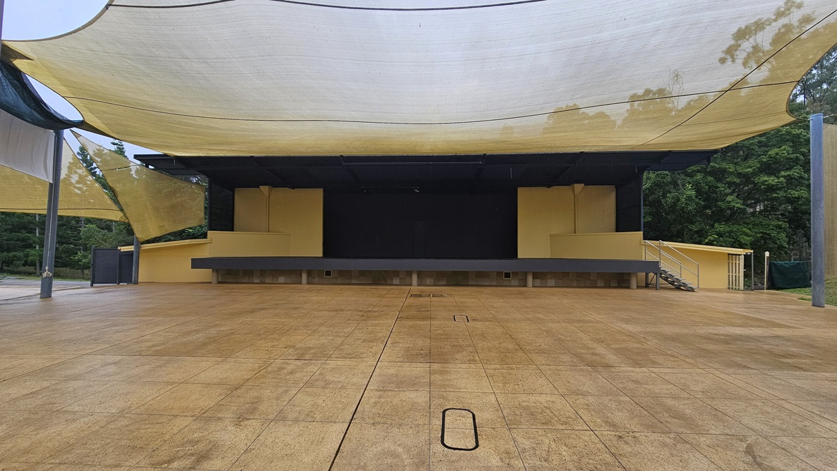 An outdoor stage with a large, flat, open area in front, covered by a series of beige fabric canopies. The stage is elevated and painted in a light yellow colour, with a black backdrop. There are steps on the right side leading up to the stage. The ground is paved with large, square tiles. Trees and greenery are visible in the background on the right side. An outdoor stage with a large, flat, open area in front, covered by a series of beige fabric canopies. The stage is elevated and painted in a light yellow colour, with a black backdrop. There are steps on the right side leading up to the stage. The ground is paved with large, square tiles. Trees and greenery are visible in the background on the right side.