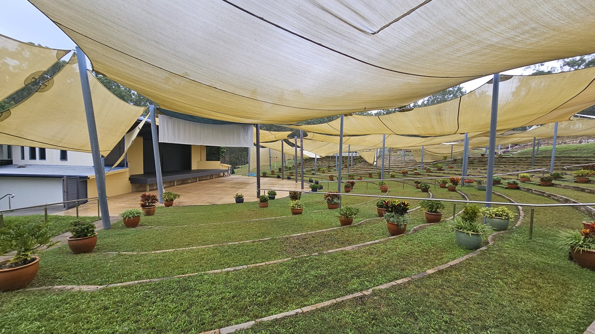 A terraced garden with neatly arranged potted plants on grassy steps. Overhead, large yellow shade cloths are stretched across metal poles, providing cover. In the background, a modern building with a sloped roof and a small stage area is visible. The ground is a mix of grass and paved paths, and the area is surrounded by a railing. A terraced garden with neatly arranged potted plants on grassy steps. Overhead, large yellow shade cloths are stretched across metal poles, providing cover. In the background, a modern building with a sloped roof and a small stage area is visible. The ground is a mix of grass and paved paths, and the area is surrounded by a railing.