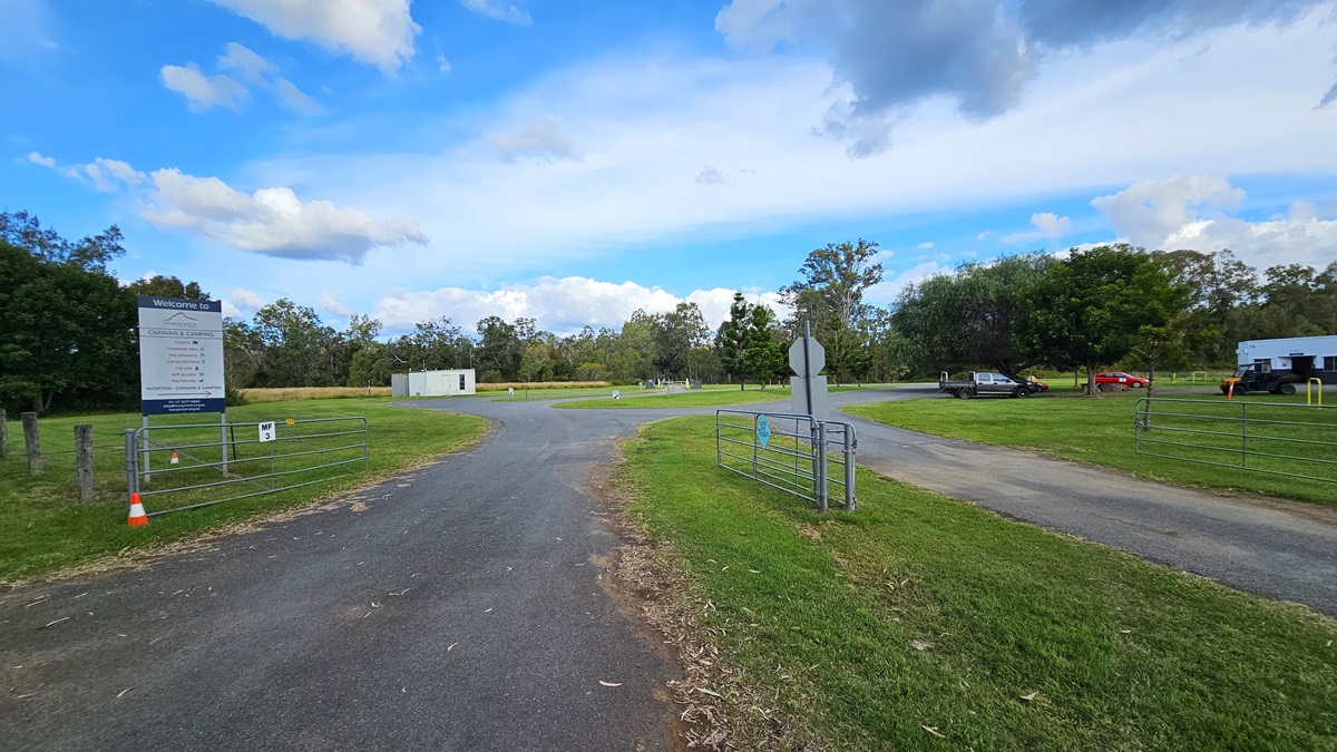 A paved road leads into a grassy area with a large sign on the left, next to a wire fence and an orange traffic cone. In the background, there are trees and a small white building. On the right, a few vehicles are parked near another building. The sky is partly cloudy. A paved road leads into a grassy area with a large sign on the left, next to a wire fence and an orange traffic cone. In the background, there are trees and a small white building. On the right, a few vehicles are parked near another building. The sky is partly cloudy.