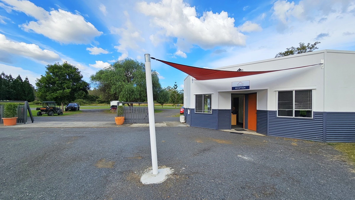 A small building with a sign reading "Reception" above the entrance is on the right. The building has a white and blue exterior with a red door. A red triangular shade sail is attached to a white pole in the foreground. On the left, there are trees and a few parked vehicles in the background. The ground is covered with gravel, and there are large potted plants near the entrance. A small building with a sign reading "Reception" above the entrance is on the right. The building has a white and blue exterior with a red door. A red triangular shade sail is attached to a white pole in the foreground. On the left, there are trees and a few parked vehicles in the background. The ground is covered with gravel, and there are large potted plants near the entrance.
