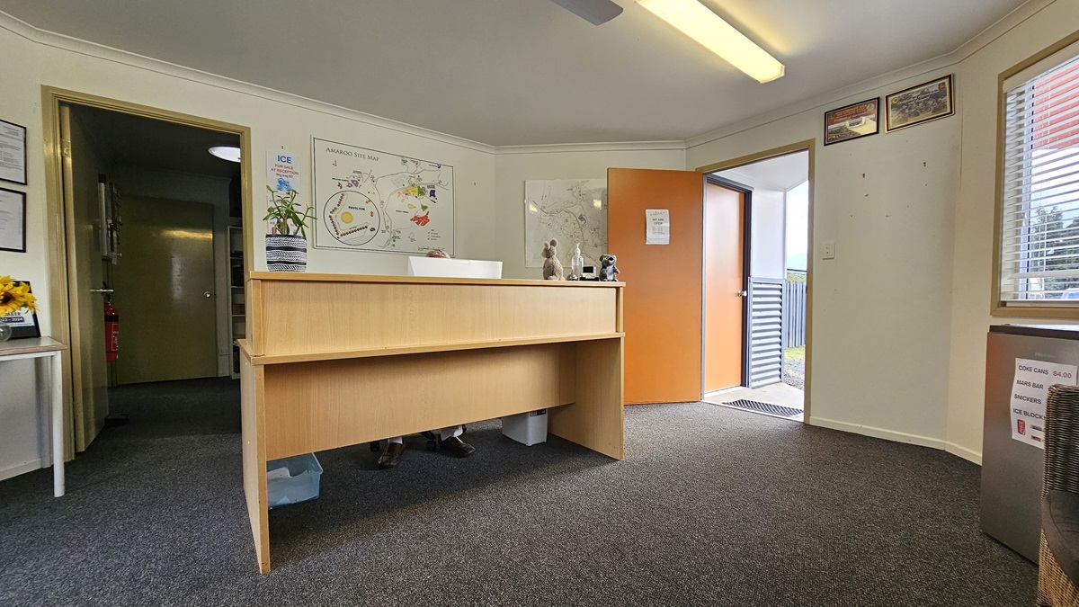 Reception area with a wooden desk in the centre. Behind the desk, a wall features a colourful map and a small plant. To the left, an open doorway leads to another room. On the right, an open door leads outside, with a view of a railing. The floor is carpeted in grey, and the ceiling has a fluorescent light fixture. A window with blinds is on the right wall, and framed certificates are visible on the left wall. Reception area with a wooden desk in the centre. Behind the desk, a wall features a colourful map and a small plant. To the left, an open doorway leads to another room. On the right, an open door leads outside, with a view of a railing. The floor is carpeted in grey, and the ceiling has a fluorescent light fixture. A window with blinds is on the right wall, and framed certificates are visible on the left wall.