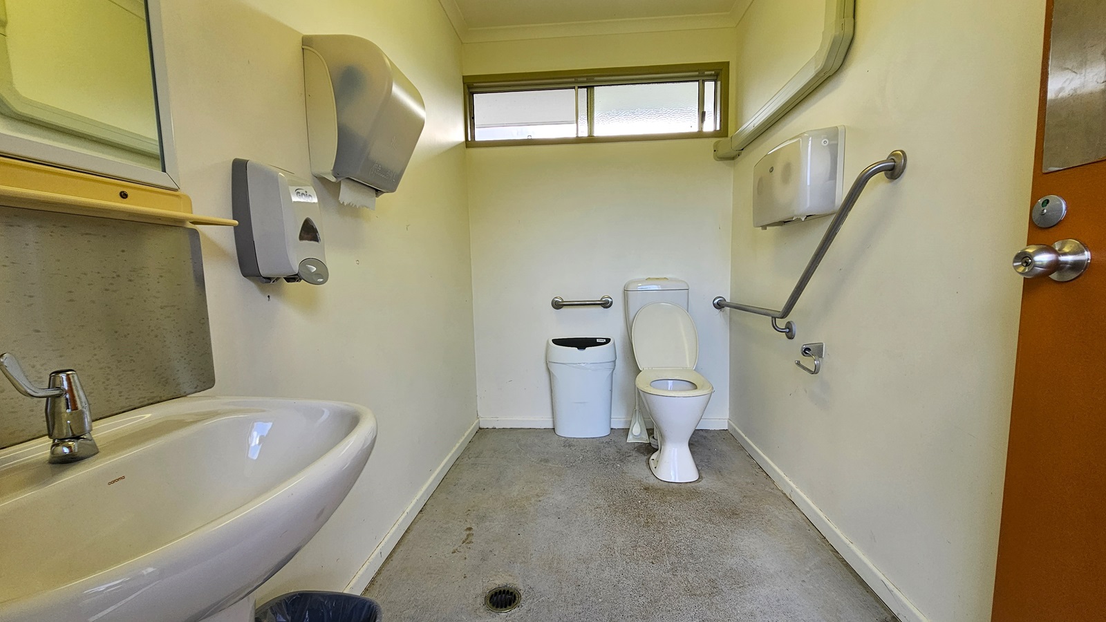 A small bathroom with a white toilet and a bin in the centre. On the left, there's a sink with a mirror above it, and a soap dispenser and hand dryer mounted on the wall. On the right, a grab rail is attached to the wall near the toilet, with a toilet paper holder beneath it. The floor is grey and slightly stained, and a narrow window is positioned high on the back wall. The door on the right has a metal handle. A small bathroom with a white toilet and a bin in the centre. On the left, there's a sink with a mirror above it, and a soap dispenser and hand dryer mounted on the wall. On the right, a grab rail is attached to the wall near the toilet, with a toilet paper holder beneath it. The floor is grey and slightly stained, and a narrow window is positioned high on the back wall. The door on the right has a metal handle.