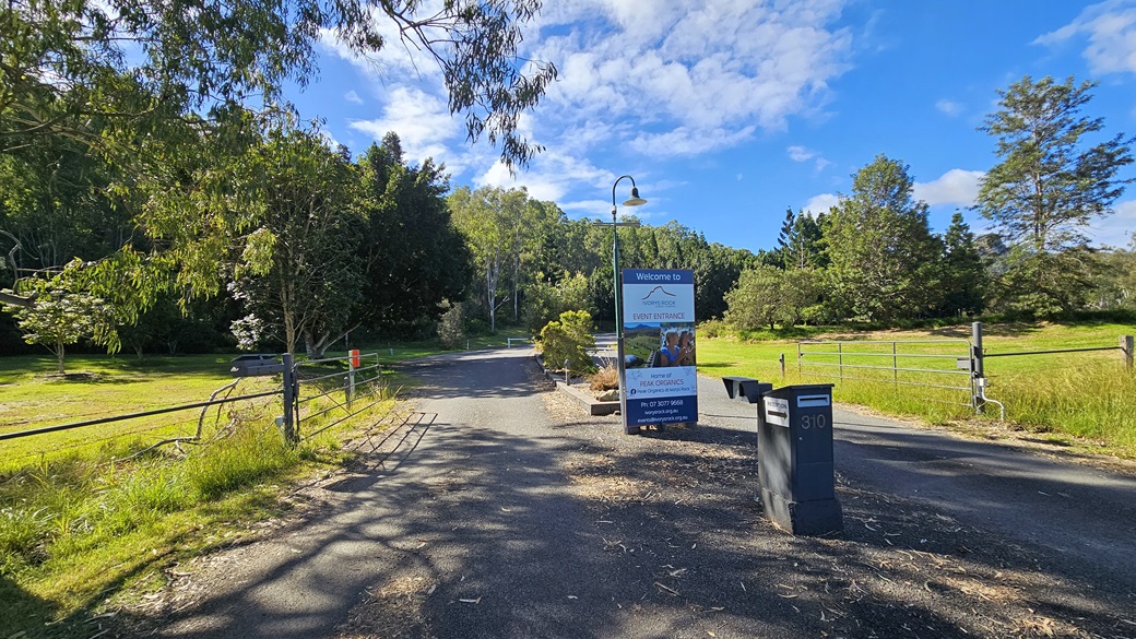 A rural road with a metal gate on the left and a grassy field on the right. In the centre, there's a signpost with a lamp above it, displaying information. Two letterboxes are positioned on the right side of the road. The background features dense trees and greenery under a partly cloudy sky. The ground is paved with some scattered leaves and debris. A rural road with a metal gate on the left and a grassy field on the right. In the centre, there's a signpost with a lamp above it, displaying information. Two letterboxes are positioned on the right side of the road. The background features dense trees and greenery under a partly cloudy sky. The ground is paved with some scattered leaves and debris.