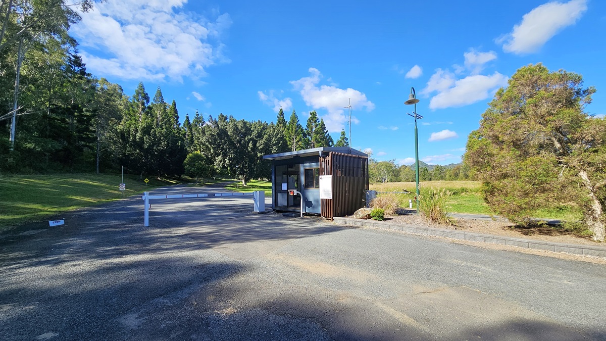 A small wooden booth is situated on the right side of a paved road, with a barrier gate in front. To the left, there is a grassy area with tall trees in the background. On the right, a lamp post stands near some bushes and a tree. The road is bordered by greenery, and the sky is clear with a few clouds. A small wooden booth is situated on the right side of a paved road, with a barrier gate in front. To the left, there is a grassy area with tall trees in the background. On the right, a lamp post stands near some bushes and a tree. The road is bordered by greenery, and the sky is clear with a few clouds.