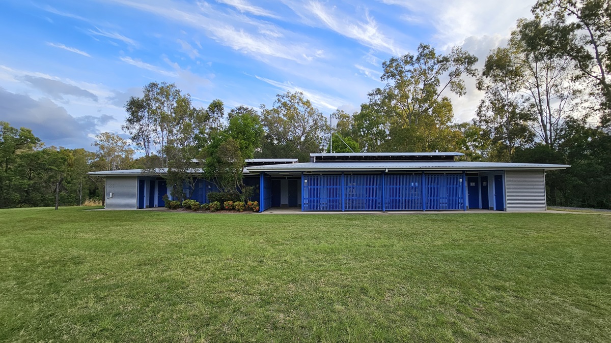 A modern, single-storey building with a flat roof and blue vertical slats on the facade. Solar panels are visible on the roof. The structure is surrounded by a well-maintained grassy lawn. Trees and shrubs are in the background, providing a natural setting. The building has a minimalist design with a focus on clean lines and functionality. A modern, single-storey building with a flat roof and blue vertical slats on the facade. Solar panels are visible on the roof. The structure is surrounded by a well-maintained grassy lawn. Trees and shrubs are in the background, providing a natural setting. The building has a minimalist design with a focus on clean lines and functionality.