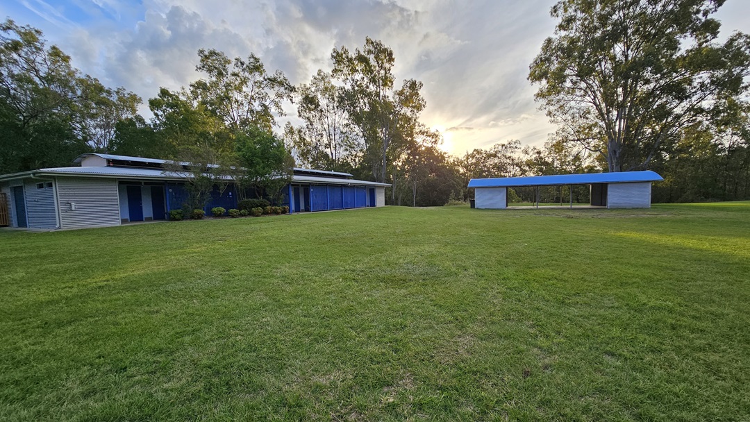 A grassy field with two structures. On the left, a long building with a flat roof and blue doors, surrounded by trees. On the right, a smaller open shelter with a blue roof. The background features tall trees and a setting sun. A grassy field with two structures. On the left, a long building with a flat roof and blue doors, surrounded by trees. On the right, a smaller open shelter with a blue roof. The background features tall trees and a setting sun.