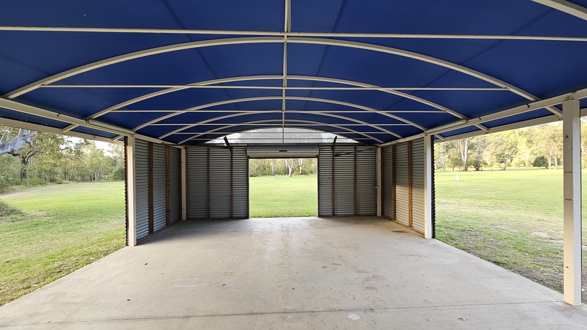 A large, open-sided shelter with a blue arched roof supported by a metal frame. The floor is concrete, and the walls are made of corrugated metal. The structure opens out to a grassy field with trees in the background. The ceiling features a series of curved beams. The left and right sides of the shelter are open, providing a view of the surrounding greenery. A large, open-sided shelter with a blue arched roof supported by a metal frame. The floor is concrete, and the walls are made of corrugated metal. The structure opens out to a grassy field with trees in the background. The ceiling features a series of curved beams. The left and right sides of the shelter are open, providing a view of the surrounding greenery.