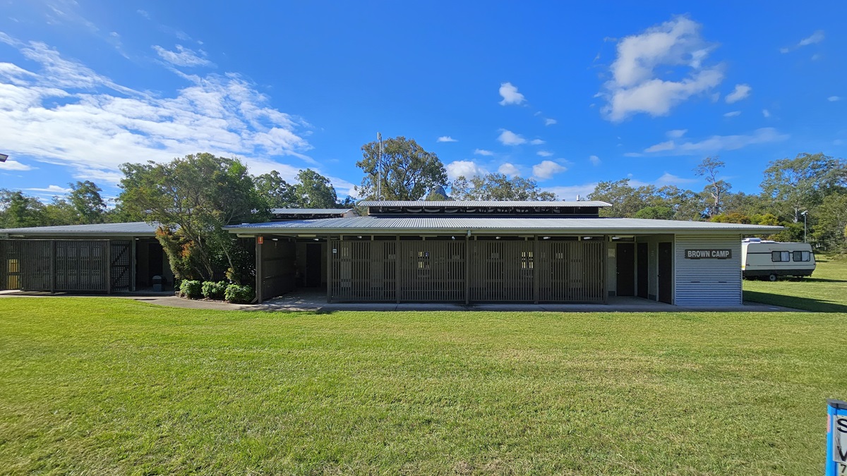 A single-storey building with a flat roof and wooden slatted exterior. On the right, there's a sign that reads "Showers & Toilets." The building is surrounded by a well-maintained grassy area. In the background, there are trees and a caravan parked to the right. The sky is clear with a few clouds. A single-storey building with a flat roof and wooden slatted exterior. On the right, there's a sign that reads "Showers & Toilets." The building is surrounded by a well-maintained grassy area. In the background, there are trees and a caravan parked to the right. The sky is clear with a few clouds.