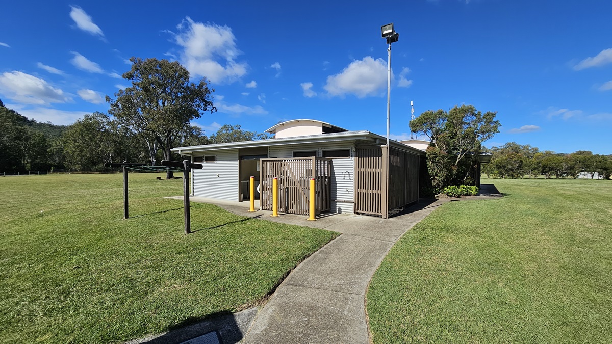 A small, single-storey building with a corrugated metal exterior and wooden slats on the front. Three yellow bollards stand in front of the entrance. A tall light pole is positioned to the right. The building is surrounded by a large grassy field with a few trees in the background. A concrete path leads up to the building, and there is a clothesline to the left. The sky is clear with a few clouds. A small, single-storey building with a corrugated metal exterior and wooden slats on the front. Three yellow bollards stand in front of the entrance. A tall light pole is positioned to the right. The building is surrounded by a large grassy field with a few trees in the background. A concrete path leads up to the building, and there is a clothesline to the left. The sky is clear with a few clouds.