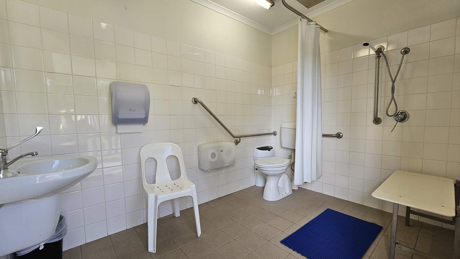 A bathroom with white tiled walls and a beige tiled floor. On the left, there's a sink with a lever tap and a wall-mounted paper towel dispenser. A white plastic chair is next to the sink. In the centre, a toilet with a grab rail beside it. On the right, a shower area with a handheld showerhead and another grab rail, separated by a white curtain. A blue mat is on the floor near the shower, and a small bench is positioned in the shower area. A bathroom with white tiled walls and a beige tiled floor. On the left, there's a sink with a lever tap and a wall-mounted paper towel dispenser. A white plastic chair is next to the sink. In the centre, a toilet with a grab rail beside it. On the right, a shower area with a handheld showerhead and another grab rail, separated by a white curtain. A blue mat is on the floor near the shower, and a small bench is positioned in the shower area.