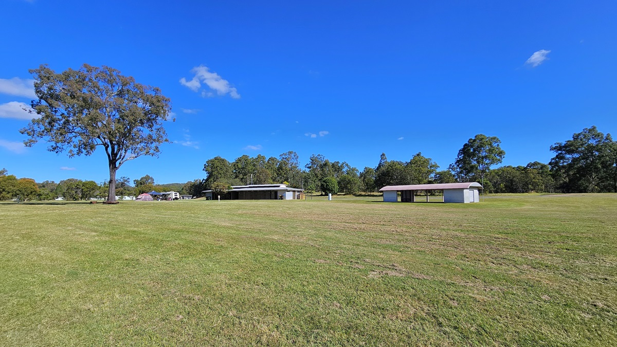 A large grassy field with a single tall tree on the left. In the middle distance, there are two small, simple buildings with flat roofs. The background is lined with dense trees. The sky is clear and blue with a few small clouds. A large grassy field with a single tall tree on the left. In the middle distance, there are two small, simple buildings with flat roofs. The background is lined with dense trees. The sky is clear and blue with a few small clouds.