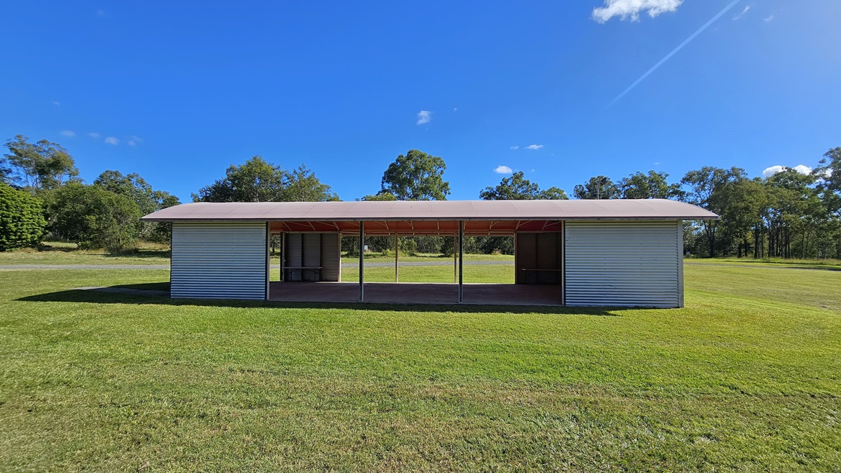A small, rectangular structure with a corrugated metal exterior stands in the centre of a grassy field. The building has an open front with a roof supported by several vertical beams. Trees and shrubs are visible in the background, surrounding the field. The grass is neatly trimmed, and the sky is clear with a few clouds. A small, rectangular structure with a corrugated metal exterior stands in the centre of a grassy field. The building has an open front with a roof supported by several vertical beams. Trees and shrubs are visible in the background, surrounding the field. The grass is neatly trimmed, and the sky is clear with a few clouds.