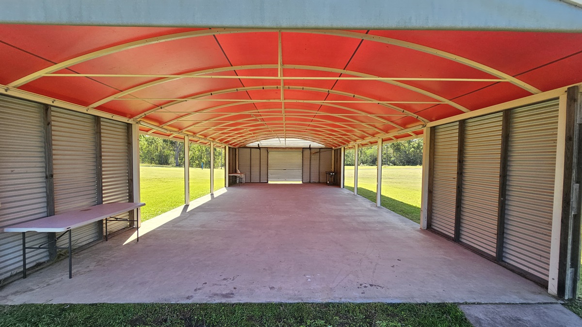 A long, open-sided structure with a red arched roof and a concrete floor. On the left, there is a folding table against a wall of closed metal shutters. The right side also features metal shutters. The background shows a grassy field and trees visible through the open sides. The ceiling is supported by metal beams. A long, open-sided structure with a red arched roof and a concrete floor. On the left, there is a folding table against a wall of closed metal shutters. The right side also features metal shutters. The background shows a grassy field and trees visible through the open sides. The ceiling is supported by metal beams.