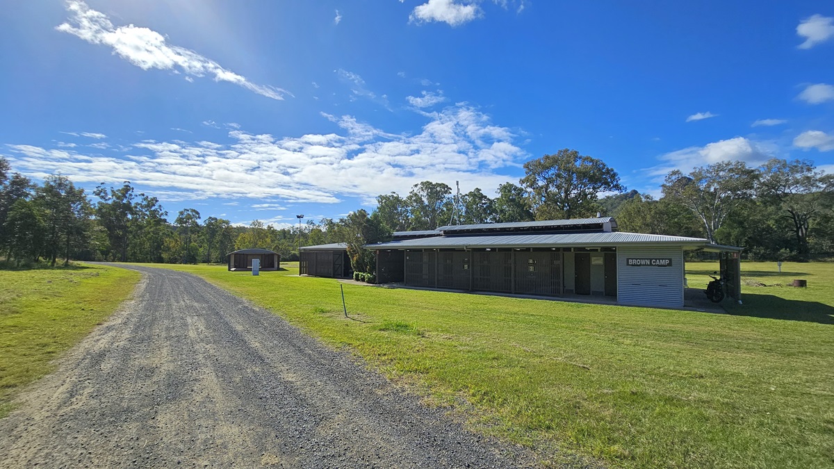 A gravel path runs from the foreground to the background on the left side, bordered by green grass. On the right, there is a long, single-storey building with a corrugated metal roof and a sign that reads "SHOWER BLOCK". Behind the building, there are trees and more greenery. The sky is mostly clear with some clouds. A gravel path runs from the foreground to the background on the left side, bordered by green grass. On the right, there is a long, single-storey building with a corrugated metal roof and a sign that reads "SHOWER BLOCK". Behind the building, there are trees and more greenery. The sky is mostly clear with some clouds.