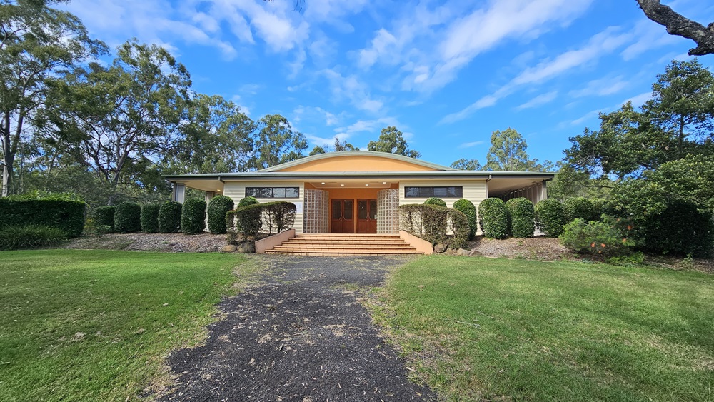 A single-storey house with a wide, sloping roof and a central entrance featuring wooden double doors. The house is surrounded by neatly trimmed hedges and bushes. A paved path leads up to the entrance, flanked by green lawns on either side. Tall trees are visible in the background, framing the house. A single-storey house with a wide, sloping roof and a central entrance featuring wooden double doors. The house is surrounded by neatly trimmed hedges and bushes. A paved path leads up to the entrance, flanked by green lawns on either side. Tall trees are visible in the background, framing the house.