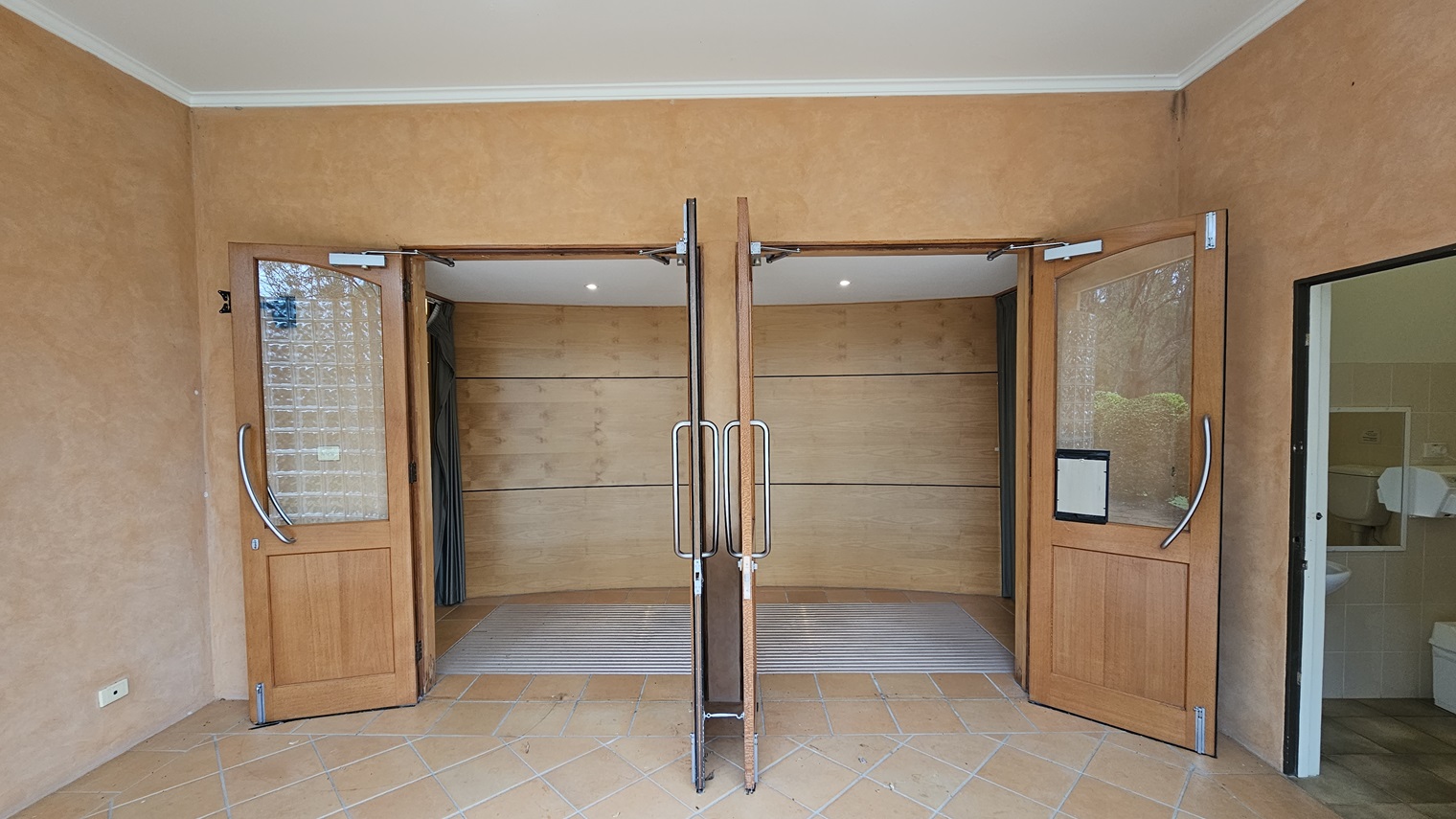 Two wooden doors with glass panels are open, leading into a room with a curved wall. The doors have metal handles. The floor is tiled in a light brown colour. On the right, there is a partially visible room with a sink and a toilet. The walls are a warm beige colour, and the ceiling is white with recessed lighting. Two wooden doors with glass panels are open, leading into a room with a curved wall. The doors have metal handles. The floor is tiled in a light brown colour. On the right, there is a partially visible room with a sink and a toilet. The walls are a warm beige colour, and the ceiling is white with recessed lighting.