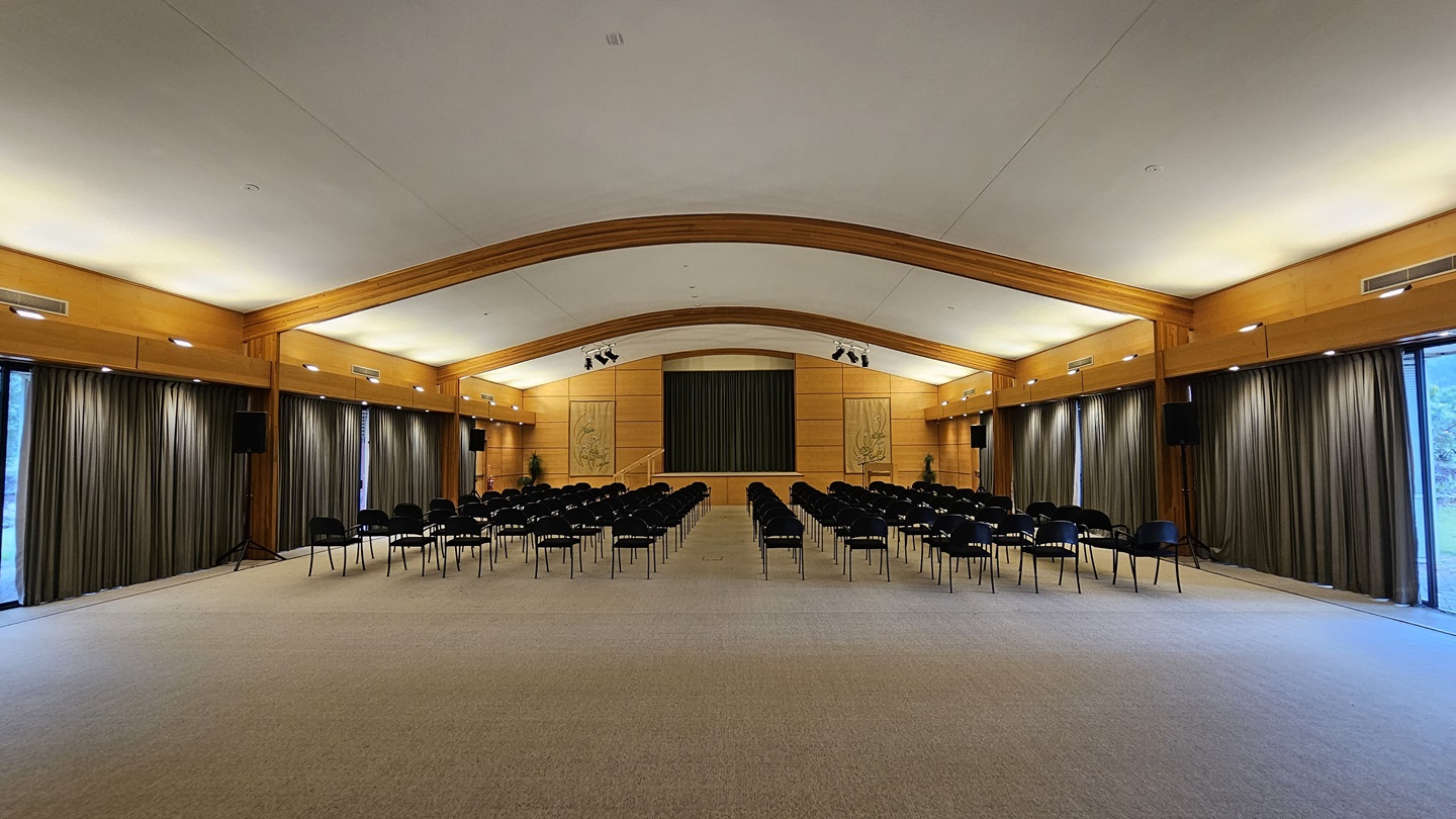 A spacious auditorium with a high, curved ceiling and wooden beams. Rows of black chairs are arranged facing a stage at the far end, which is flanked by two large decorative panels. The walls are lined with tall, dark curtains, and there are speakers mounted on the walls. The floor is carpeted in a light colour, and the room is well-lit with overhead lighting. A spacious auditorium with a high, curved ceiling and wooden beams. Rows of black chairs are arranged facing a stage at the far end, which is flanked by two large decorative panels. The walls are lined with tall, dark curtains, and there are speakers mounted on the walls. The floor is carpeted in a light colour, and the room is well-lit with overhead lighting.