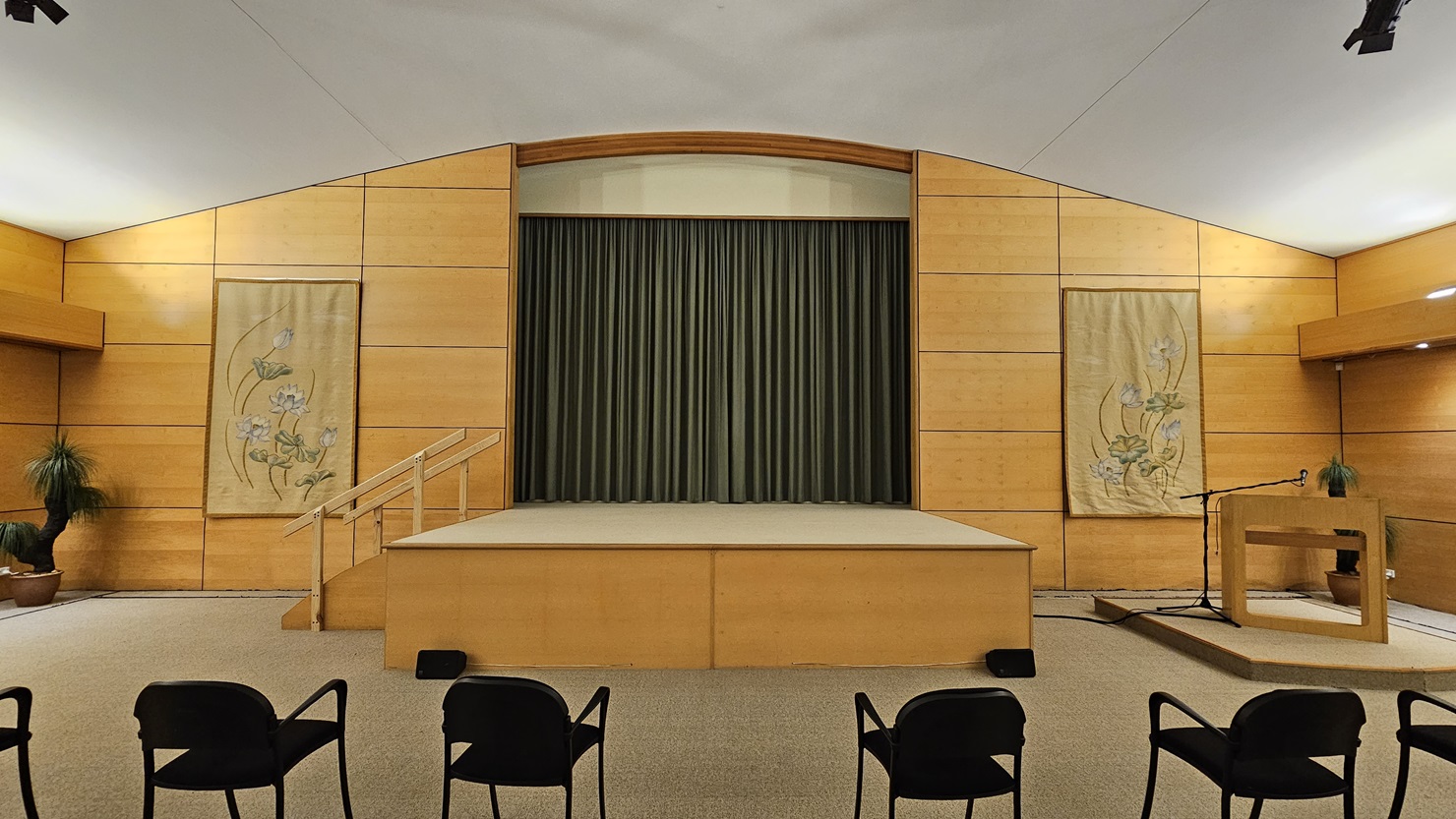 A room with a stage in the centre, featuring a green curtain backdrop. The walls are wooden with two decorative panels on either side of the stage, each displaying floral designs. On the right, there's a lectern with a microphone and a small plant. Several black chairs are positioned in the foreground. The ceiling is white, and the floor is carpeted. A potted plant is visible in the left corner. A room with a stage in the centre, featuring a green curtain backdrop. The walls are wooden with two decorative panels on either side of the stage, each displaying floral designs. On the right, there's a lectern with a microphone and a small plant. Several black chairs are positioned in the foreground. The ceiling is white, and the floor is carpeted. A potted plant is visible in the left corner.