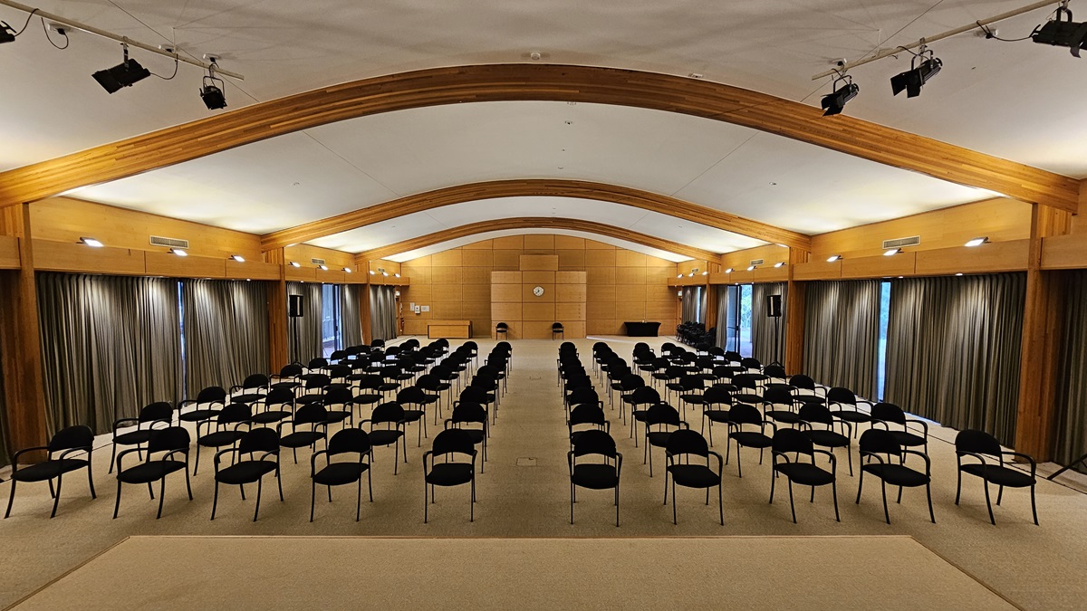 A large conference room with rows of black chairs arranged in the centre, facing a wooden panelled wall at the back. The ceiling is arched with wooden beams, and the walls are lined with long, dark curtains. The floor is carpeted in a light colour. Overhead, there are several spotlights attached to the ceiling beams. A clock is mounted on the back wall, and there are two microphones on stands at the front. A large conference room with rows of black chairs arranged in the centre, facing a wooden panelled wall at the back. The ceiling is arched with wooden beams, and the walls are lined with long, dark curtains. The floor is carpeted in a light colour. Overhead, there are several spotlights attached to the ceiling beams. A clock is mounted on the back wall, and there are two microphones on stands at the front.