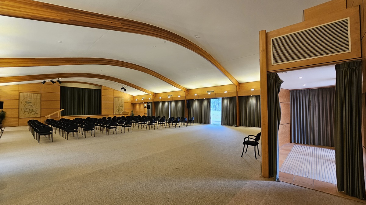 A spacious conference room with a high, curved ceiling featuring wooden beams. On the left, rows of black chairs face a stage with a green curtain. The walls are wooden with decorative panels. On the right, a doorway with dark curtains leads to another room. The floor is carpeted in a light colour, and the room is well-lit with recessed lighting along the walls. A spacious conference room with a high, curved ceiling featuring wooden beams. On the left, rows of black chairs face a stage with a green curtain. The walls are wooden with decorative panels. On the right, a doorway with dark curtains leads to another room. The floor is carpeted in a light colour, and the room is well-lit with recessed lighting along the walls.