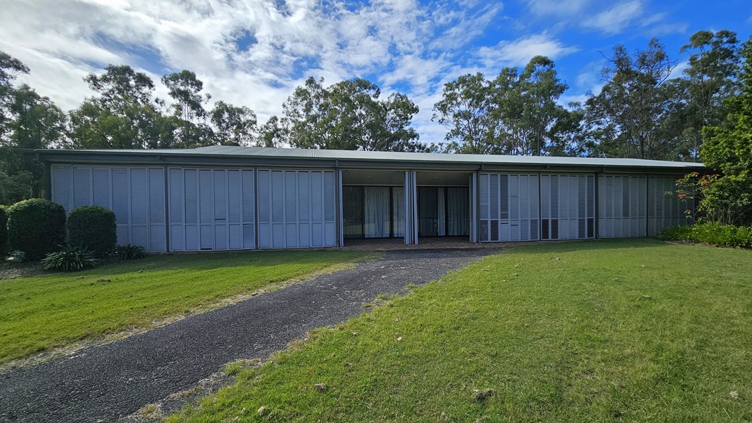 A single-storey building with a flat roof and large, vertical metal panels covering the front. A gravel path leads up to the entrance, flanked by green lawns on either side. Bushes and trees are visible on the left and right, with taller trees in the background. The sky is partly cloudy. A single-storey building with a flat roof and large, vertical metal panels covering the front. A gravel path leads up to the entrance, flanked by green lawns on either side. Bushes and trees are visible on the left and right, with taller trees in the background. The sky is partly cloudy.