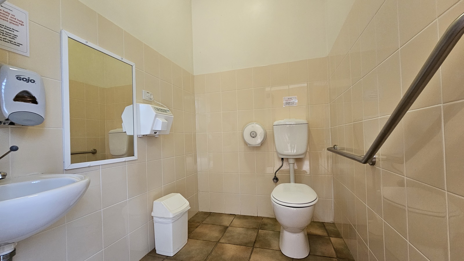 A public restroom with beige tiled walls and a brown tiled floor. On the left, there's a small white sink with a soap dispenser above it and a mirror. A paper towel dispenser is mounted on the wall. In the centre, a white toilet with a wall-mounted cistern and a toilet paper holder. On the right, a metal grab bar is attached to the wall. A small white bin is on the floor near the sink. The ceiling is plain and light-coloured. A public restroom with beige tiled walls and a brown tiled floor. On the left, there's a small white sink with a soap dispenser above it and a mirror. A paper towel dispenser is mounted on the wall. In the centre, a white toilet with a wall-mounted cistern and a toilet paper holder. On the right, a metal grab bar is attached to the wall. A small white bin is on the floor near the sink. The ceiling is plain and light-coloured.