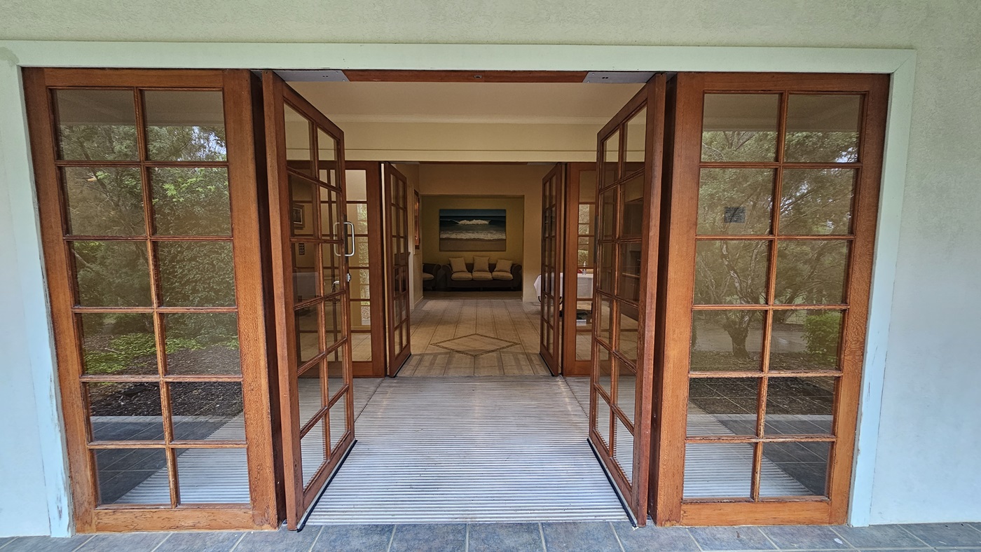 Wooden-framed glass doors open inward, leading to an interior space. The floor is tiled with a light-coloured mat at the entrance. Inside, a beige sofa with cushions is visible, set against a wall with a large painting above it. The ceiling is plain and light-coloured. On either side of the doors, there are windows showing greenery outside. Wooden-framed glass doors open inward, leading to an interior space. The floor is tiled with a light-coloured mat at the entrance. Inside, a beige sofa with cushions is visible, set against a wall with a large painting above it. The ceiling is plain and light-coloured. On either side of the doors, there are windows showing greenery outside.