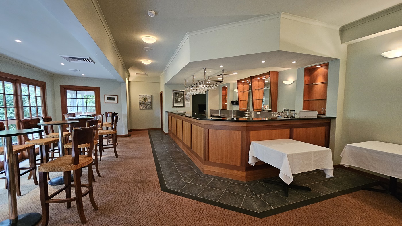 A bar area with a wooden counter on the right, featuring hanging glasses and shelves with bottles. Two tables with white tablecloths are in front of the bar. On the left, there are tall tables and chairs near large windows with wooden frames. The floor is carpeted, and the ceiling has recessed lighting. Walls are light-coloured with framed pictures. A bar area with a wooden counter on the right, featuring hanging glasses and shelves with bottles. Two tables with white tablecloths are in front of the bar. On the left, there are tall tables and chairs near large windows with wooden frames. The floor is carpeted, and the ceiling has recessed lighting. Walls are light-coloured with framed pictures.
