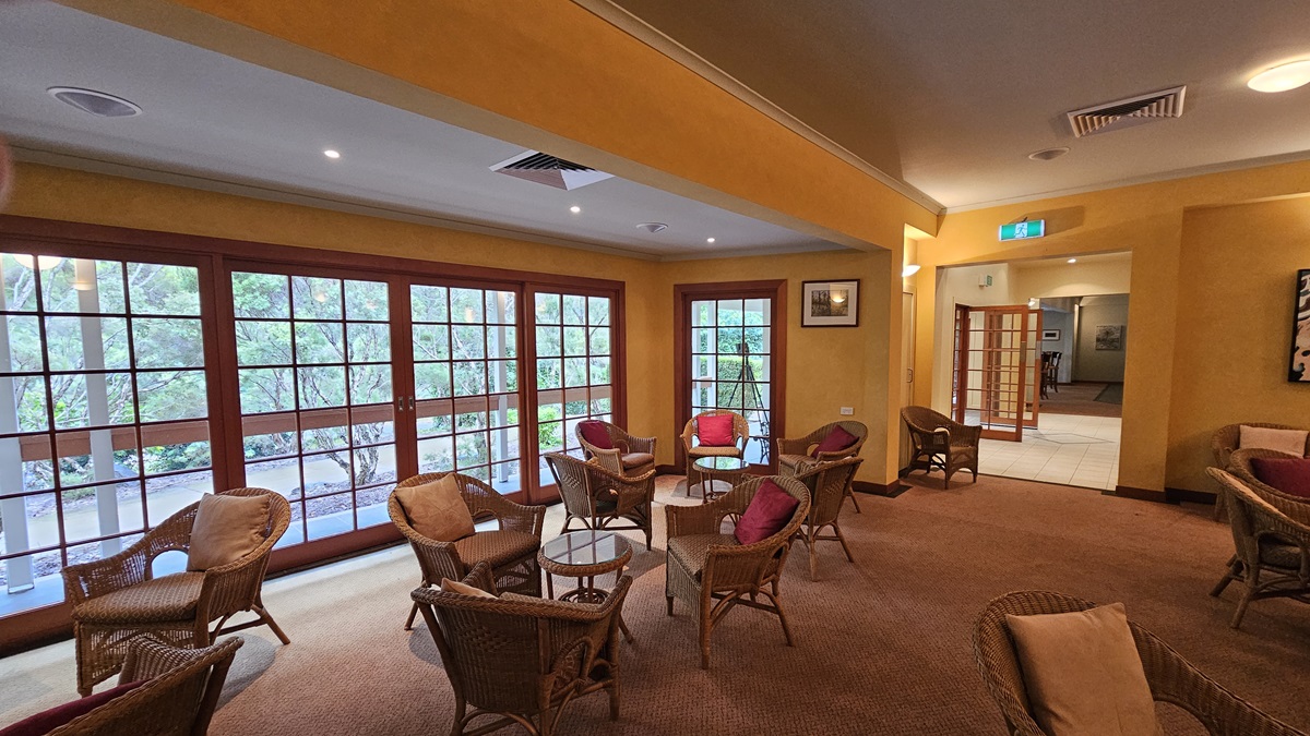 A cosy lounge area with wicker chairs and tables, each adorned with red cushions, arranged on a brown carpet. Large wooden-framed windows on the left offer a view of greenery outside. The walls are painted yellow, and the ceiling features recessed lighting and air vents. A doorway on the right leads to another room, with an exit sign above. A framed picture hangs on the wall near the doorway. A cosy lounge area with wicker chairs and tables, each adorned with red cushions, arranged on a brown carpet. Large wooden-framed windows on the left offer a view of greenery outside. The walls are painted yellow, and the ceiling features recessed lighting and air vents. A doorway on the right leads to another room, with an exit sign above. A framed picture hangs on the wall near the doorway.
