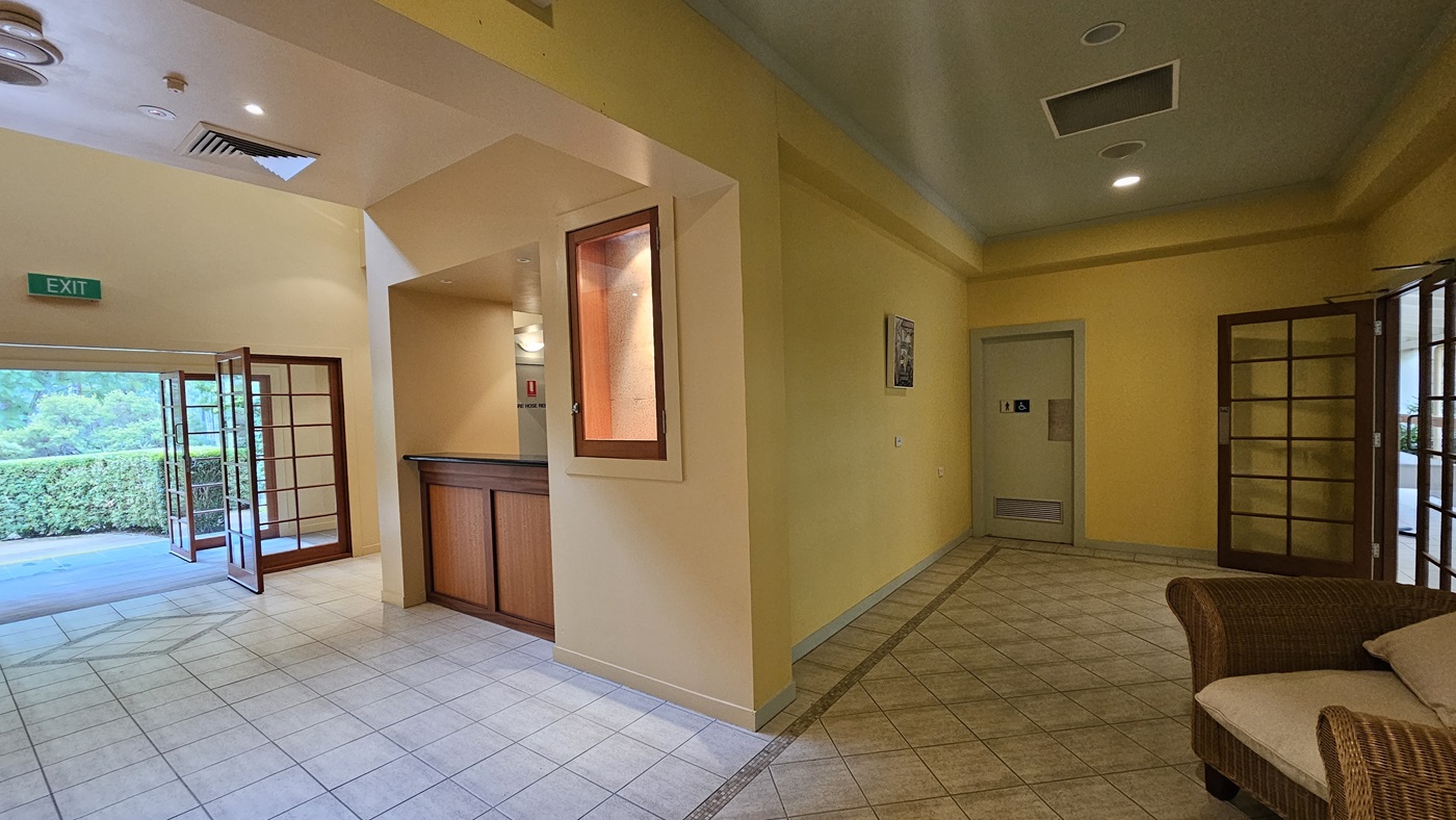 A hallway with yellow walls and a tiled floor. On the left, there is an open wooden-framed glass door leading outside, with an "EXIT" sign above. A wooden counter is visible near the centre, with a small window above it. On the right, a wicker chair with a cushion is placed against the wall. Further down the hallway, a door with a sign is visible. The ceiling has recessed lighting and ventilation panels. A hallway with yellow walls and a tiled floor. On the left, there is an open wooden-framed glass door leading outside, with an "EXIT" sign above. A wooden counter is visible near the centre, with a small window above it. On the right, a wicker chair with a cushion is placed against the wall. Further down the hallway, a door with a sign is visible. The ceiling has recessed lighting and ventilation panels.