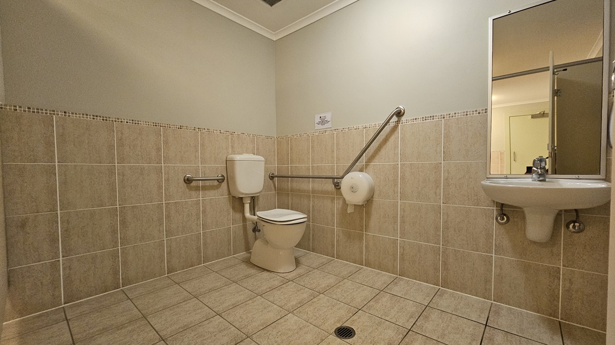 A bathroom with beige tiled walls and floor. On the left, there is a toilet with a white cistern, equipped with horizontal and diagonal metal grab bars. A toilet paper holder is attached to the diagonal bar. On the right, a white sink with a mirror above it is visible. The ceiling is plain with a light fixture. A bathroom with beige tiled walls and floor. On the left, there is a toilet with a white cistern, equipped with horizontal and diagonal metal grab bars. A toilet paper holder is attached to the diagonal bar. On the right, a white sink with a mirror above it is visible. The ceiling is plain with a light fixture.