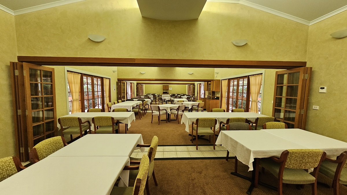 A spacious dining room with multiple rectangular tables covered in white tablecloths, surrounded by cushioned chairs. The room features large windows with wooden frames and curtains on both sides, allowing natural light to enter. The walls are painted in a warm beige tone, and there are wall-mounted lights. The floor is carpeted, and the ceiling is high with a central recessed section. A spacious dining room with multiple rectangular tables covered in white tablecloths, surrounded by cushioned chairs. The room features large windows with wooden frames and curtains on both sides, allowing natural light to enter. The walls are painted in a warm beige tone, and there are wall-mounted lights. The floor is carpeted, and the ceiling is high with a central recessed section.
