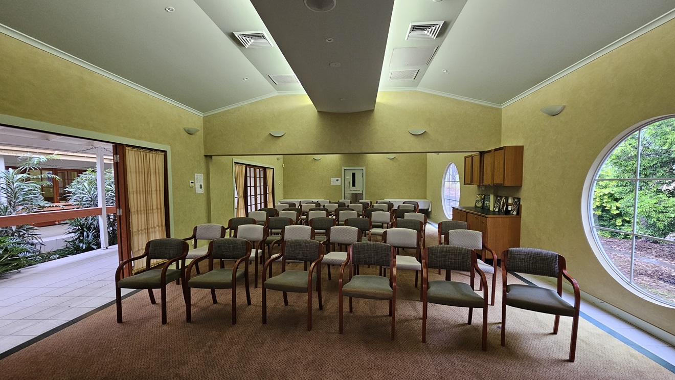 A room with rows of chairs facing forward, arranged in a semi-circle. The walls are painted yellow, and there are circular windows on the right side, offering a view of greenery outside. On the left, there is an open doorway leading to a corridor with plants visible. The ceiling is vaulted with recessed lighting and air vents. A cabinet with framed photos is on the right wall. The floor is carpeted in a muted colour. A room with rows of chairs facing forward, arranged in a semi-circle. The walls are painted yellow, and there are circular windows on the right side, offering a view of greenery outside. On the left, there is an open doorway leading to a corridor with plants visible. The ceiling is vaulted with recessed lighting and air vents. A cabinet with framed photos is on the right wall. The floor is carpeted in a muted colour.