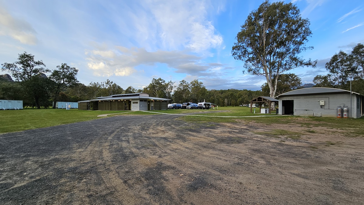 A modern, single-storey building with a flat roof and solar panels on top. The structure features wooden slats and large windows. In front, there's a well-maintained grassy area. To the left, a blue car is parked near some trees. On the right, a small shelter and more trees are visible. The background is filled with greenery and tall trees. A modern, single-storey building with a flat roof and solar panels on top. The structure features wooden slats and large windows. In front, there's a well-maintained grassy area. To the left, a blue car is parked near some trees. On the right, a small shelter and more trees are visible. The background is filled with greenery and tall trees.
