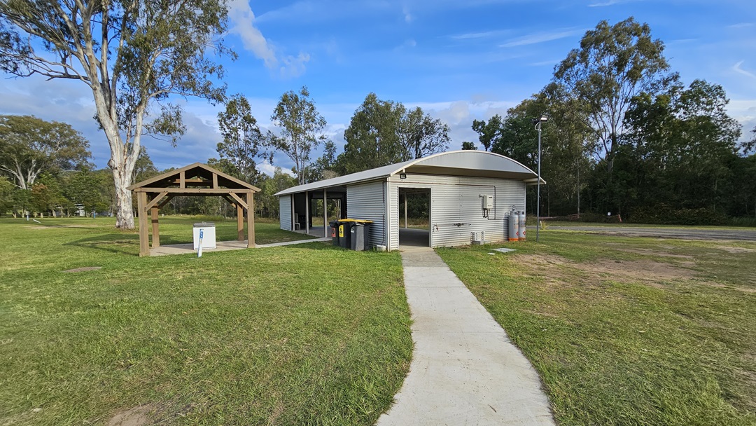 A small, corrugated metal building with a curved roof is situated on a grassy area. A concrete path leads to the entrance. To the left, there is a wooden shelter with a pitched roof and a small white utility box underneath. Trees are scattered in the background, and there are a few bins near the building. The sky is partly cloudy. A small, corrugated metal building with a curved roof is situated on a grassy area. A concrete path leads to the entrance. To the left, there is a wooden shelter with a pitched roof and a small white utility box underneath. Trees are scattered in the background, and there are a few bins near the building. The sky is partly cloudy.