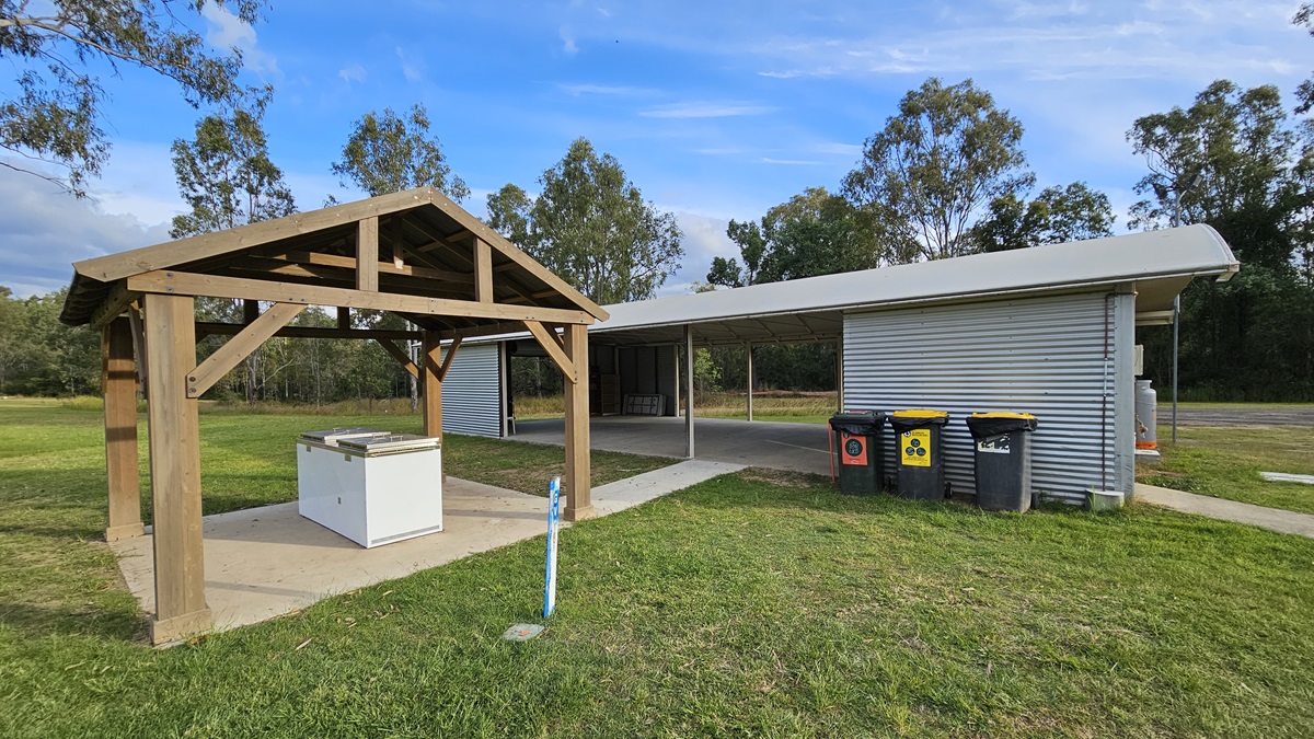 A grassy area with a wooden shelter on the left, housing a white barbecue unit on a concrete slab. To the right, a corrugated metal building with an extended roof provides shade. In front of the building are several wheelie bins in different colours. Trees are visible in the background, and the ground is covered with grass. A grassy area with a wooden shelter on the left, housing a white barbecue unit on a concrete slab. To the right, a corrugated metal building with an extended roof provides shade. In front of the building are several wheelie bins in different colours. Trees are visible in the background, and the ground is covered with grass.