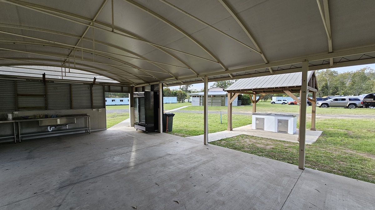 A covered outdoor area with a curved metal roof and concrete floor. On the left, there is a metal sink and counter. In the centre, a large refrigerator stands near an open doorway leading to a grassy area. On the right, a small pavilion with a metal roof and a countertop is visible. In the background, there are several parked cars and buildings, surrounded by trees. A covered outdoor area with a curved metal roof and concrete floor. On the left, there is a metal sink and counter. In the centre, a large refrigerator stands near an open doorway leading to a grassy area. On the right, a small pavilion with a metal roof and a countertop is visible. In the background, there are several parked cars and buildings, surrounded by trees.
